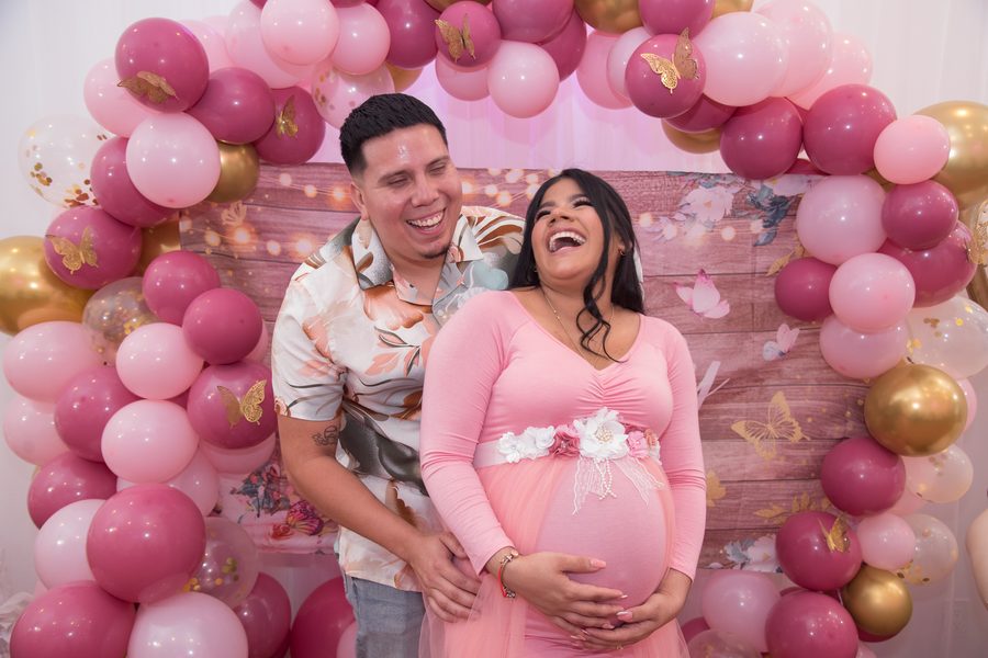 Expecting couple laughing together under a pink balloon arch at their baby shower
