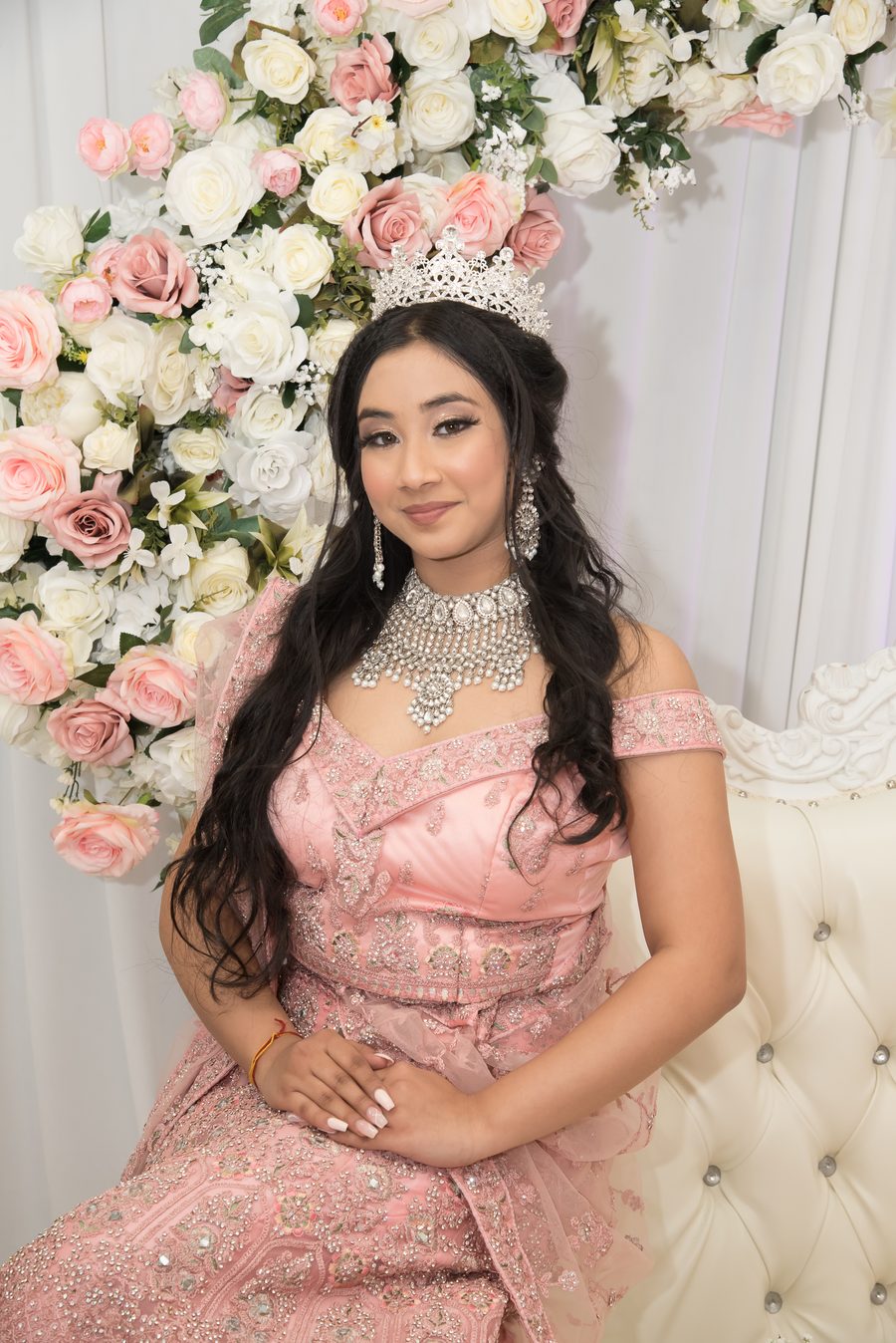 Quinceañera in pink seated before a rose wall wearing a crystal crown