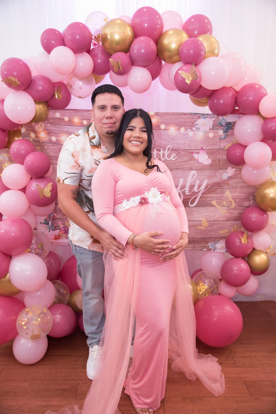Expecting couple posed beneath a pink and gold balloon arch